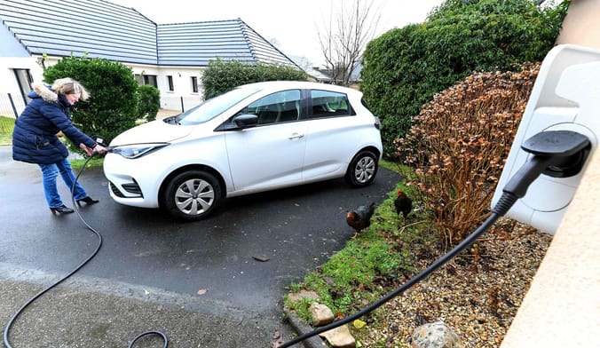 A woman in a dark blue winter coat charges a small white car in the driveway of a home