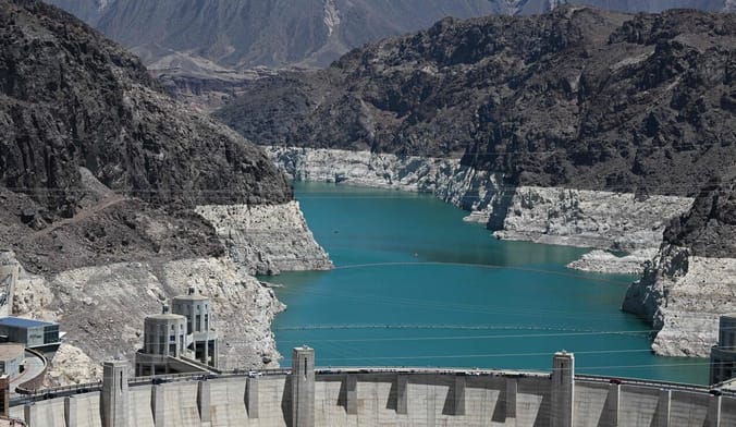 an aqua blue river at a low water level flows through a hilly landscape to a large concrete dam