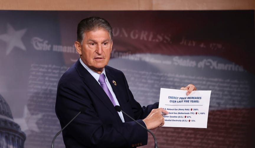 A white man in a dark blue suit and purple tie stands behind a wooden lectern and points to a paper