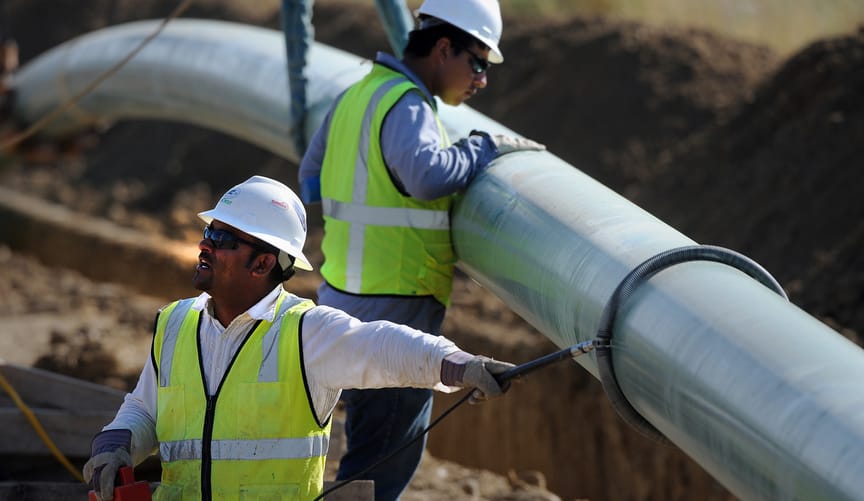 Two men in white hard hats and bright reflective vests stand near a large metal pipeline that is being lowered into a trench