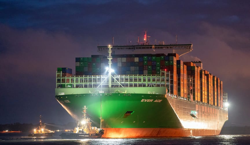A very large green cargo ship laden with cargo containers comes into port at dusk