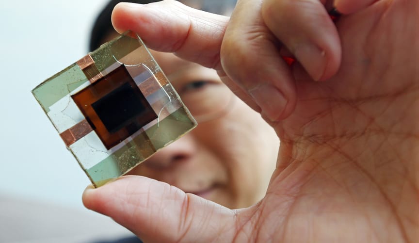 A man with black hair and wire-rimmed glasses holds a small glass square with a smaller square of dark brown material inside