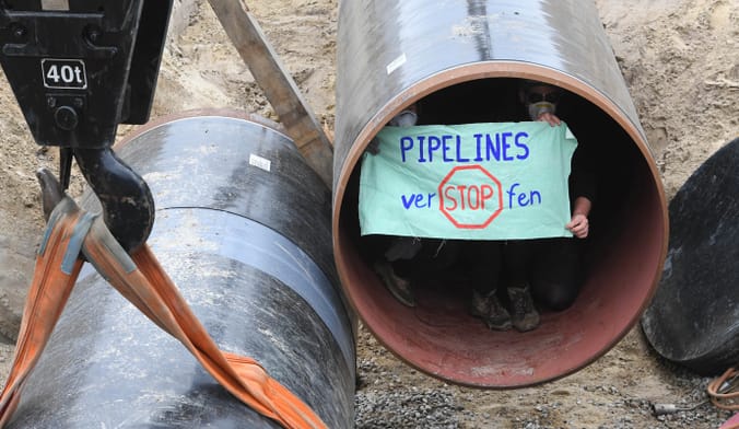 Two protestors sit inside a large section of metal pipeline holding a sign that says "Stop Pipelines"