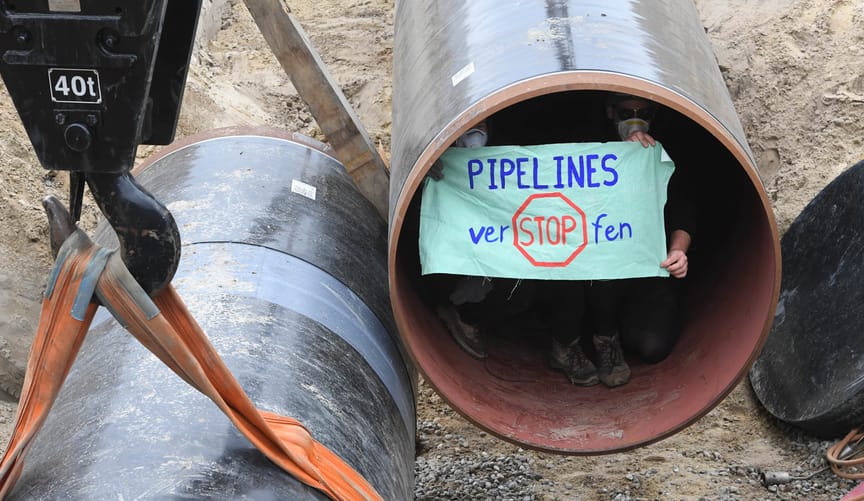 Two protestors sit inside a large section of metal pipeline holding a sign that says "Stop Pipelines"
