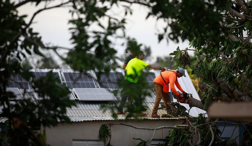 Two men in bright safety clothing remove  debris from a rooftop that also has solar panels