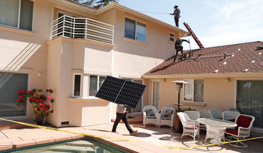 Workers install solar panels on a two-story peach stucco home with a swimming pool