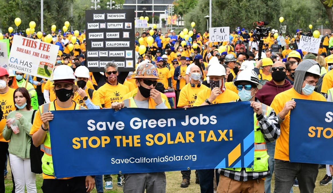 Protestors wearing masks and hard hats hold a sign that says "Save green jobs. Stop the solar tax!"