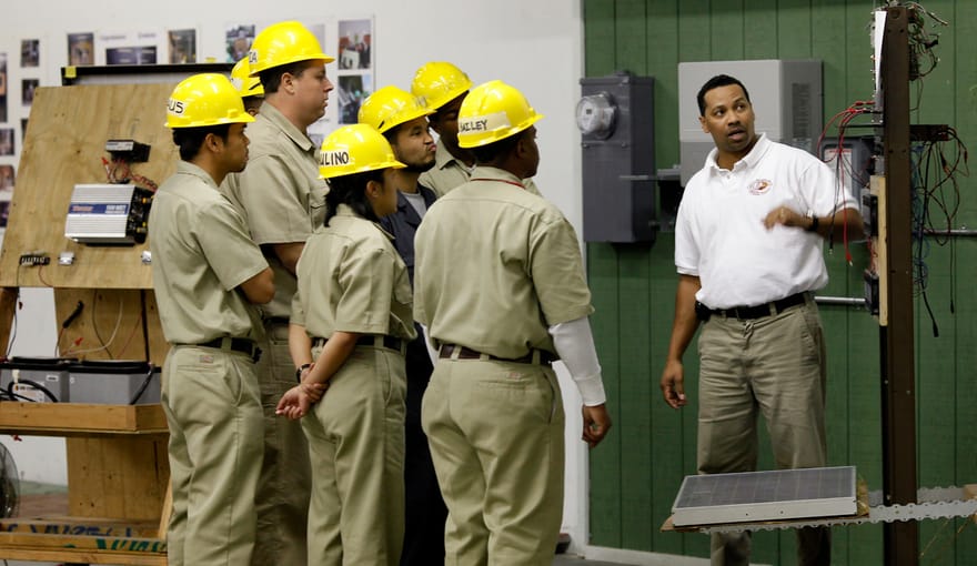 A group of people in yellow hard hats and work uniforms gather around an instructor in a training classroom