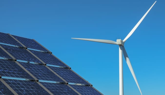 A solar panel array next to a white wind turbine against the backdrop of a bright blue sky