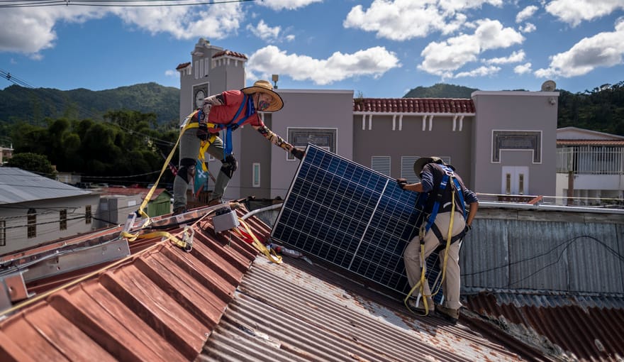 Two technicians install a blue rectangular solar panel on a rooftop with red tiles. A blue cloudy sky is overhead