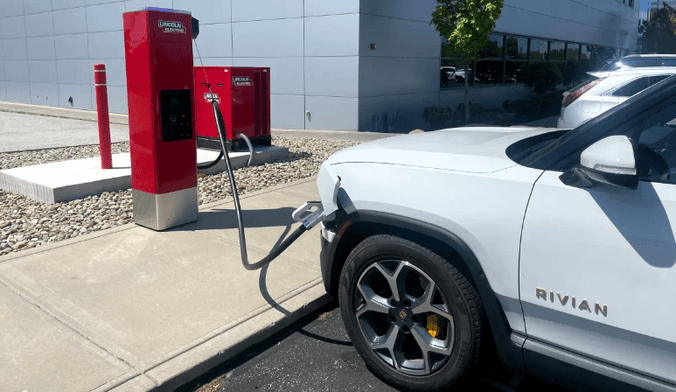 A white Rivian electric vehicle plugged into a red charger in front of a gray, multistory building