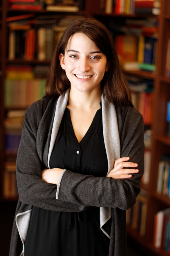 A woman with brown shoulder-length hair stands with her arms cross in front of tall bookshelves