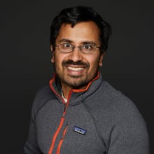 A smiling man with dark hair and glasses wearing a gray heathered pullover with orange trim