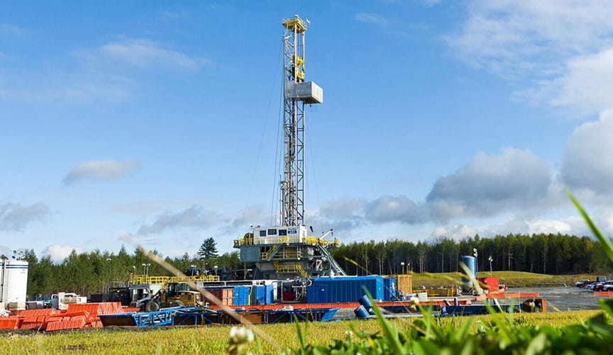 A large drilling rig is surrounded by other industrial equipment in a prairie. A grove of trees can be seen in the distance.