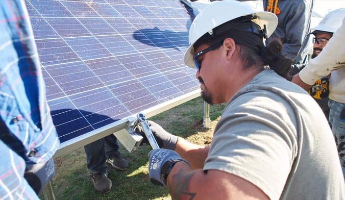 A man with black hair in a ponytail wearing a white hardhat installs a solar panel.