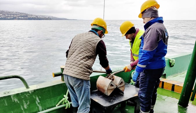 On the Black Sea, three people on a green ship deck probe some machinery that has collected sediment samples