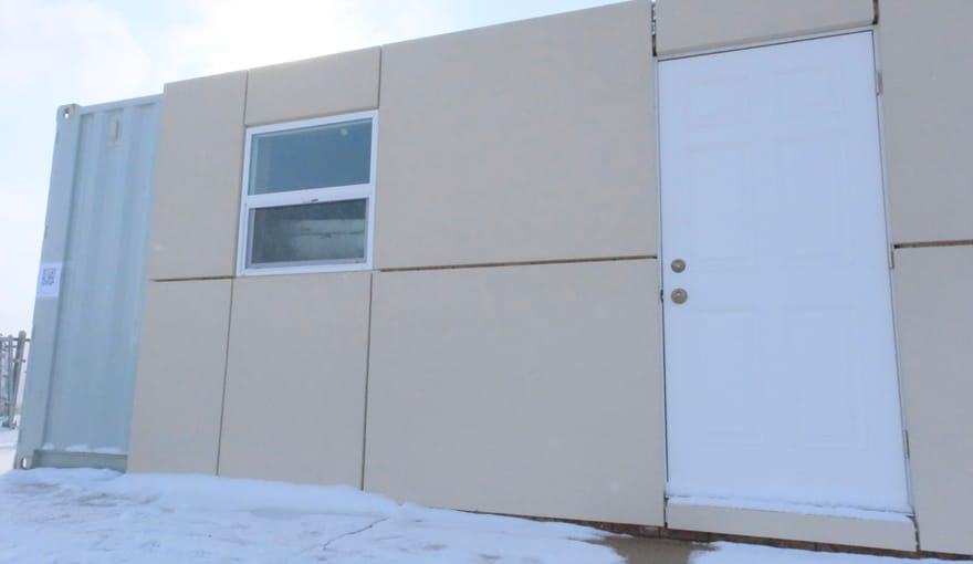 A metal shipping container with a window and door is clad in blocks of beige insulating polystyrene panels.