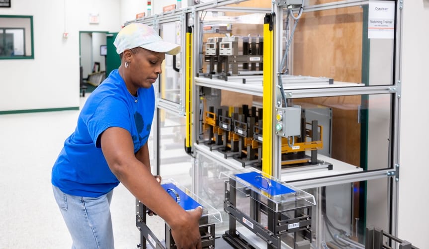 A Black woman wearing a baseball cap prepares to lift a specialized metal and plastic container.