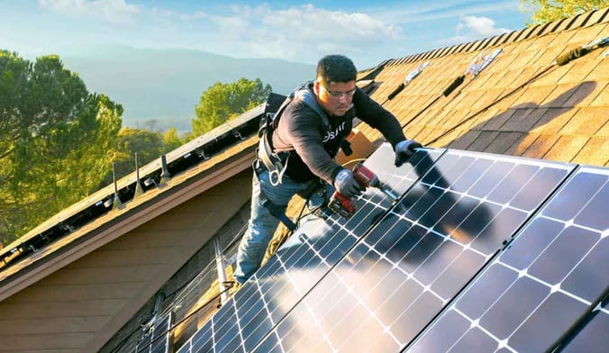 A man installs a solar panel on a rooftop with green mountains in the background