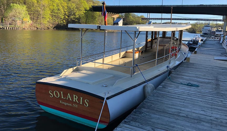 A long, narrow, open-sided boat docked on a river. The name SOLARIS is emblazoned on its back panel.