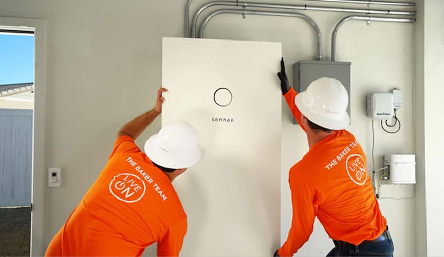 Two men in work uniforms and white hard hats install a metal rectangular white box in a home's garage