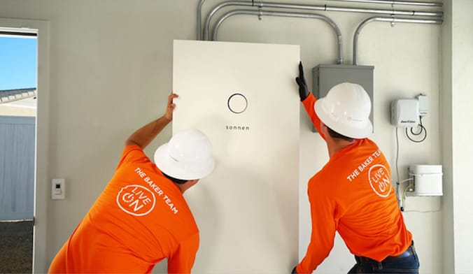 Two men in work uniforms and white hard hats install a metal rectangular white box in a home's garage