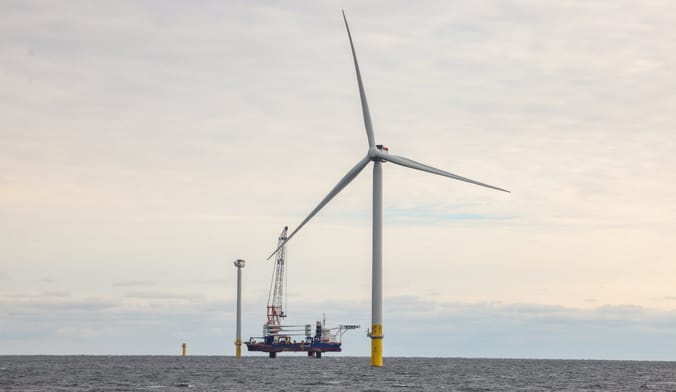 Wind turbines and a lifting barge are shown on the open ocean under a cloudy gray sky