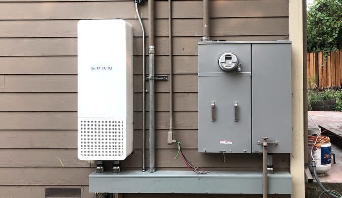 A Span smart electrical panel installed on the side of a home next to an old-fashioned electrical panel with utility meter