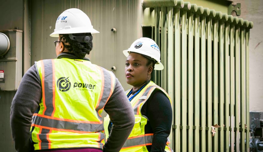 Two women in hard hats and reflective safety vests stand next to a large electrical component