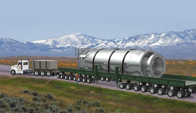 A tractor-trailer carrying a large metal object. In the background is a snowy mountain range