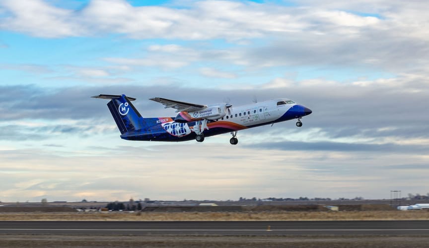 A white-and-blue airplane with twin propellers takes off from an airport landing strip.