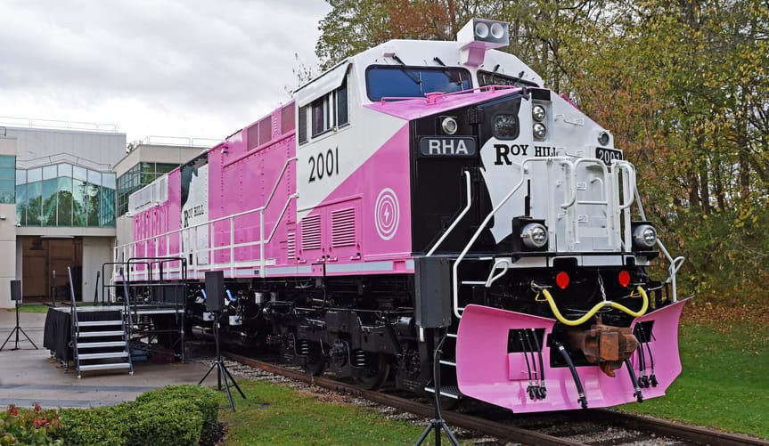 A large pink train sits on tracks in front of an office building and next to tall trees
